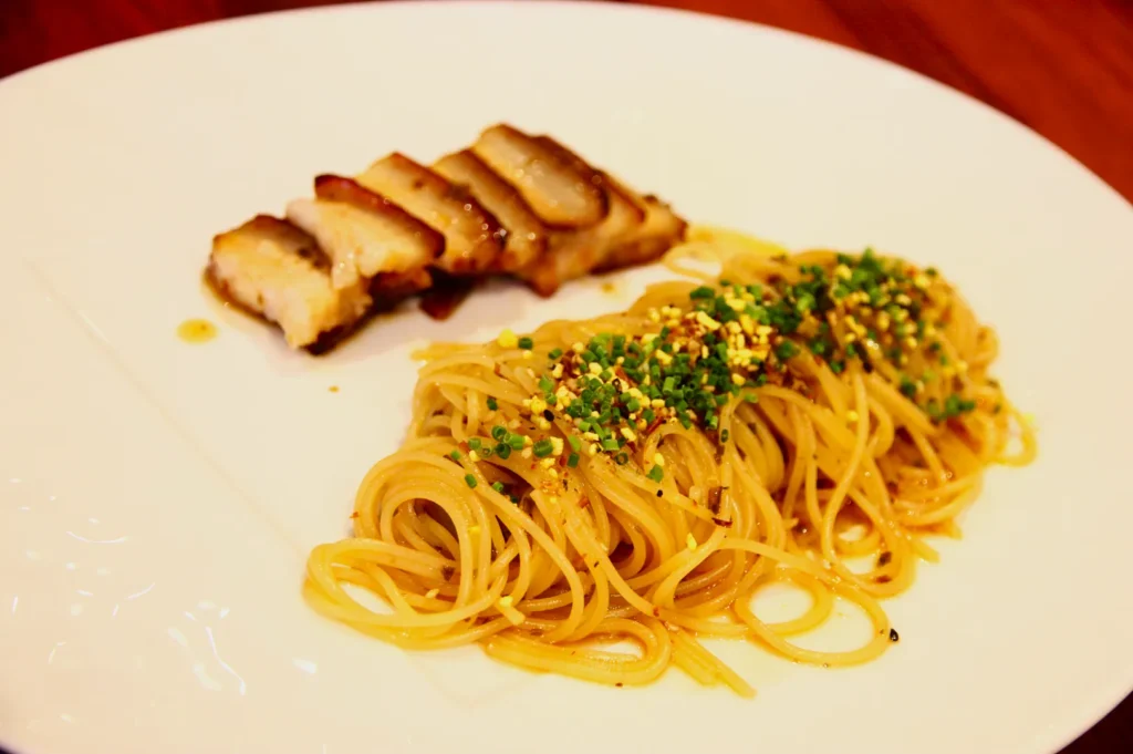 A plate with seasoned spaghetti topped with green herbs and sesame seeds, accompanied by sliced grilled pork, exuding a warm and inviting ambiance.