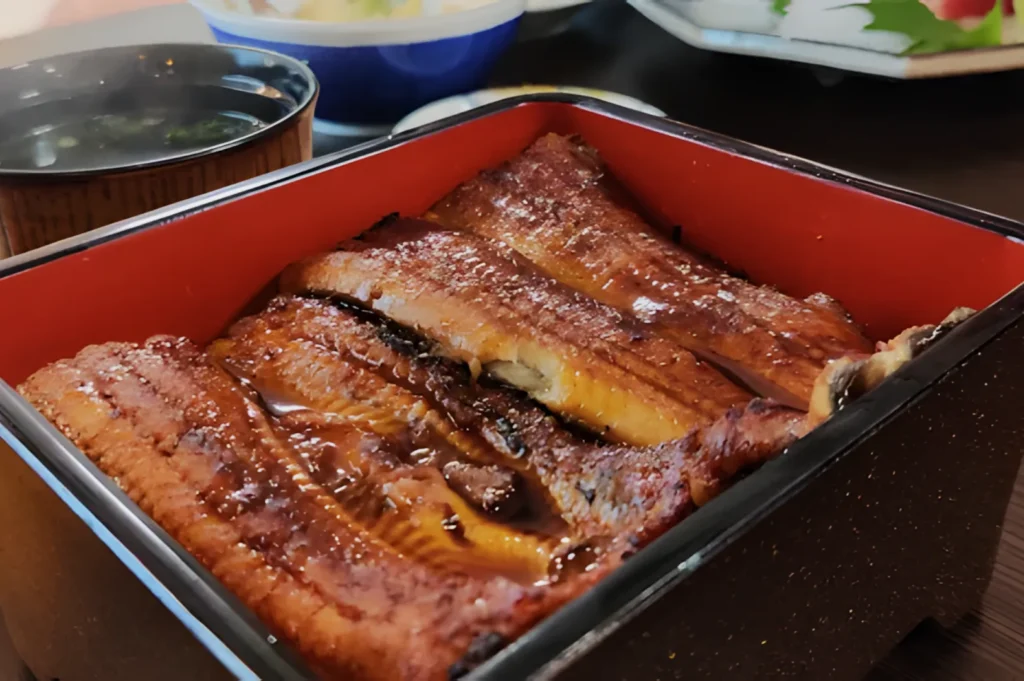 Grilled eel covered in glossy sauce, served over rice in a lacquer box. A bowl of soup is blurred in the background, creating a cozy dining atmosphere.