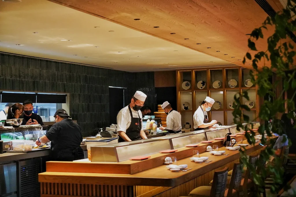 Chefs in white uniforms and hats prepare food behind a sushi bar in a warmly lit, modern restaurant. The ambiance is lively and inviting.
