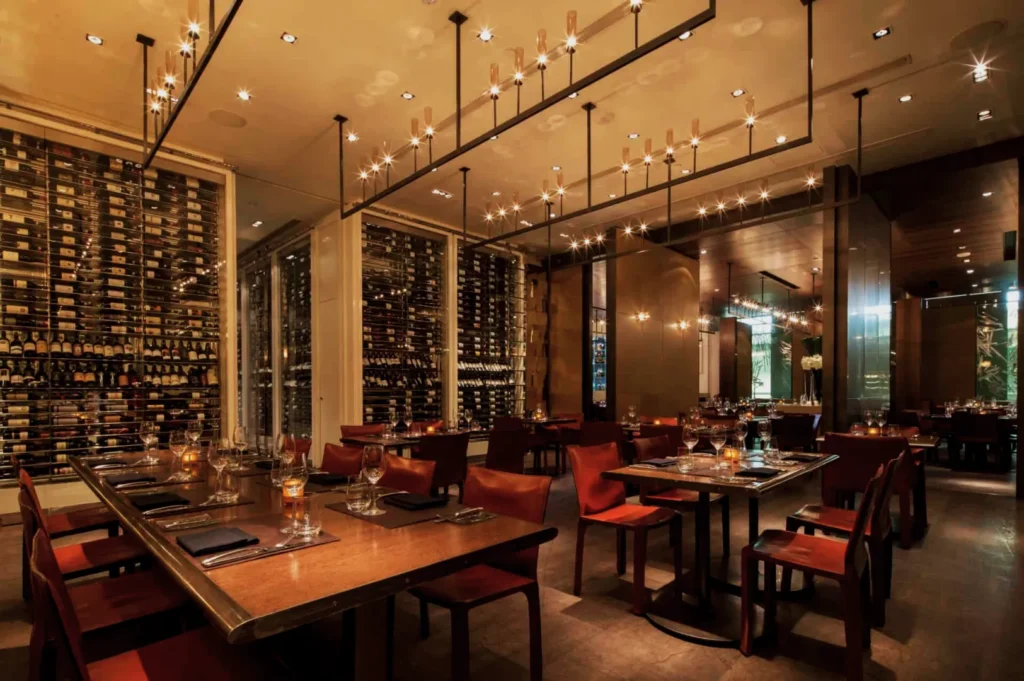 Upscale restaurant interior with warm lighting, elegant chandeliers, wine bottles lining the walls, wooden tables set with glassware, and brown leather chairs.