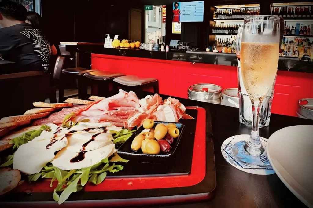 A charcuterie board with assorted meats, cheeses, and olives sits on a table beside a glass of champagne. The setting is a vibrant, modern bar with red accents