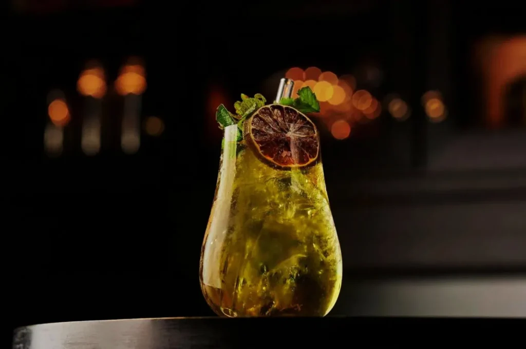 A pear-shaped glass filled with a luminous yellow cocktail, garnished with a dried lime slice and fresh mint, set against a dark, blurred background.