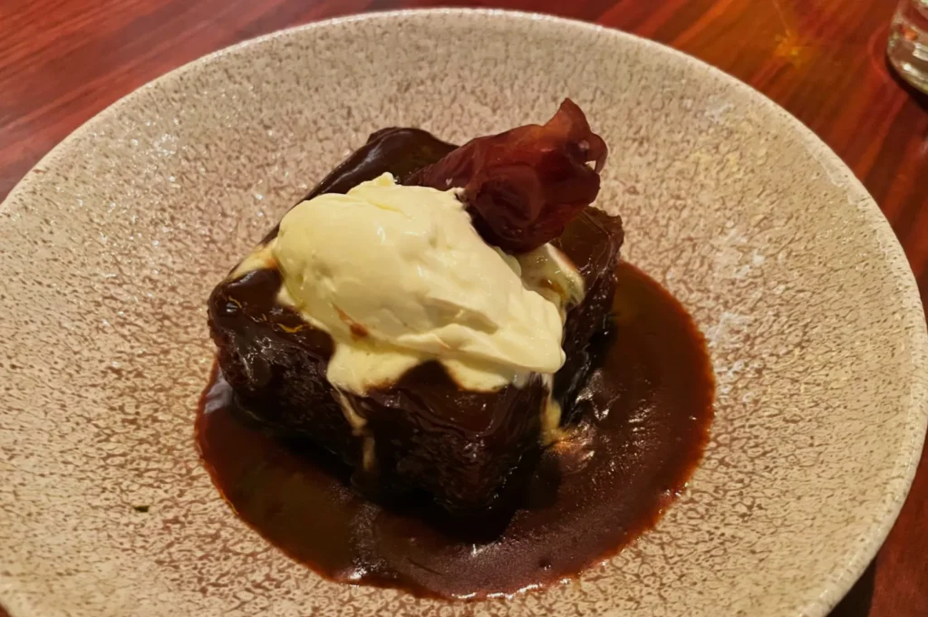 A rich, gooey chocolate brownie topped with a dollop of vanilla ice cream and a small piece of candied fruit, served in a shallow dish on a wooden table.