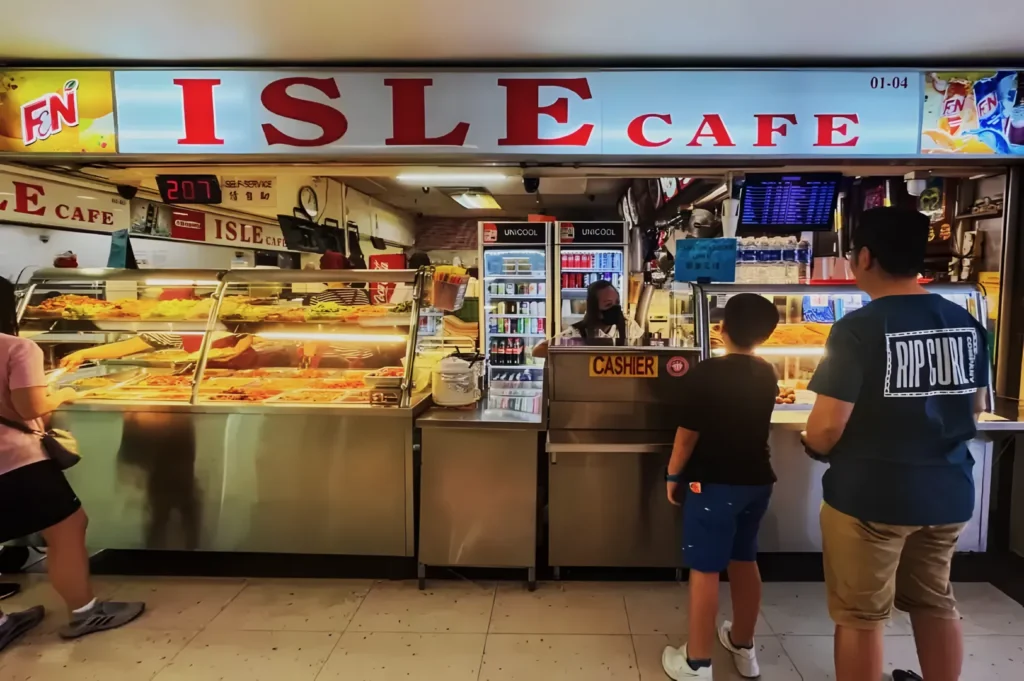 A bustling food counter at ISLE Cafe, displaying warm meals under glass. Two customers stand at the cashier, while a staff member works inside.