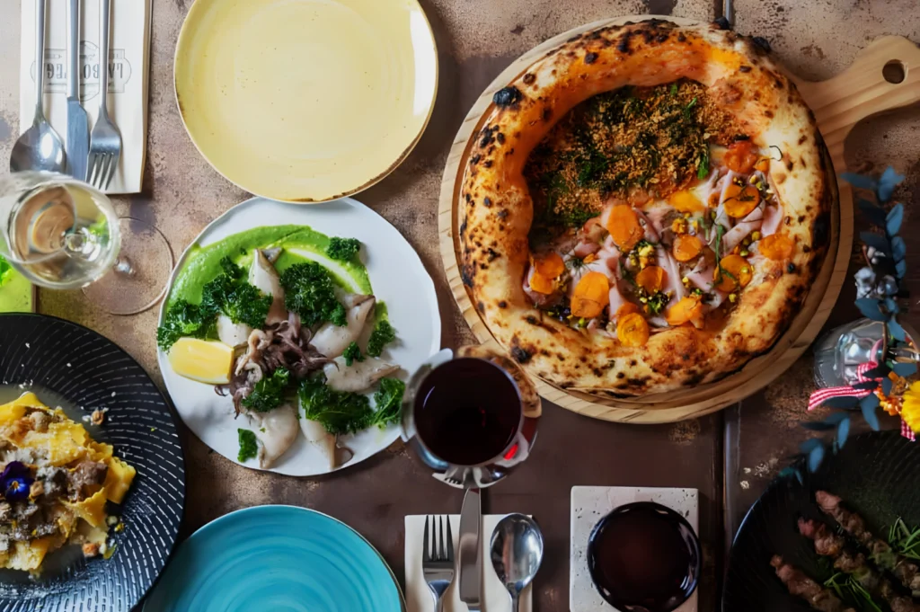 A table with a rustic pizza topped with greens and orange slices, surrounded by dishes of seafood and pasta, two glasses of wine, and utensils.