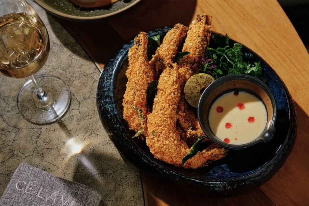 Crispy coated prawns on a dark plate with fresh greens, a lime wedge, and a creamy dipping sauce with red drops. Nearby, a glass of white wine.