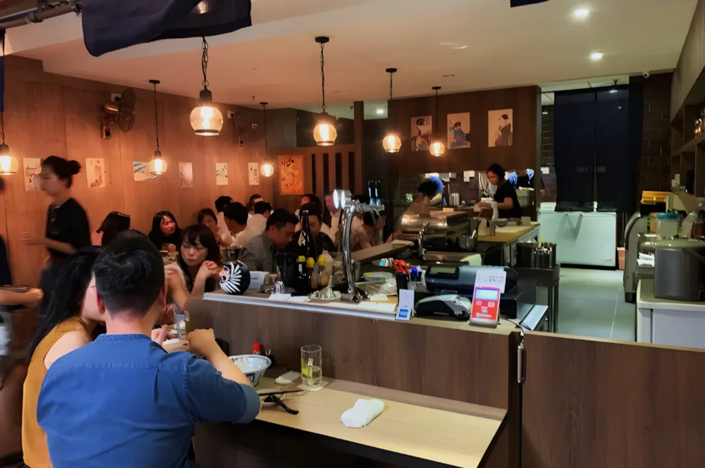 A cozy restaurant interior with patrons dining at wooden tables, lit by warm pendant lights. A busy atmosphere with servers attending to guests.