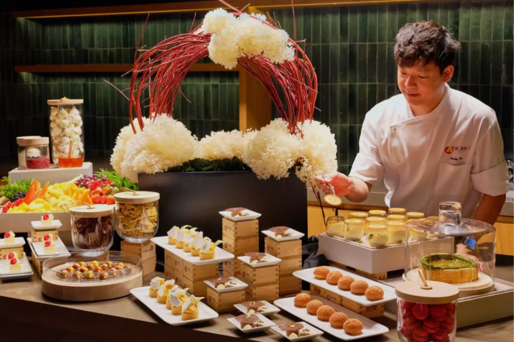 A chef in a white uniform is arranging an elegant dessert display with assorted pastries, jars, and a decorative floral arrangement on a table.