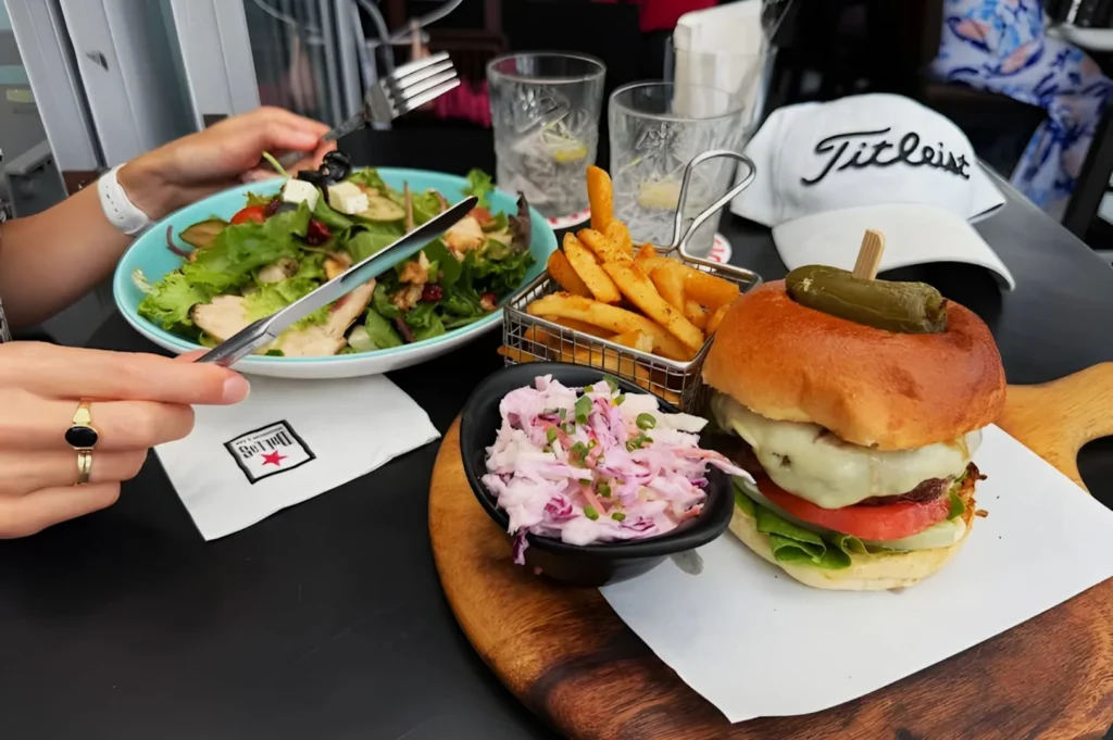 A gourmet burger with fries and coleslaw is served on a wooden platter beside a fresh salad. A white cap and glasses of water are in the background.