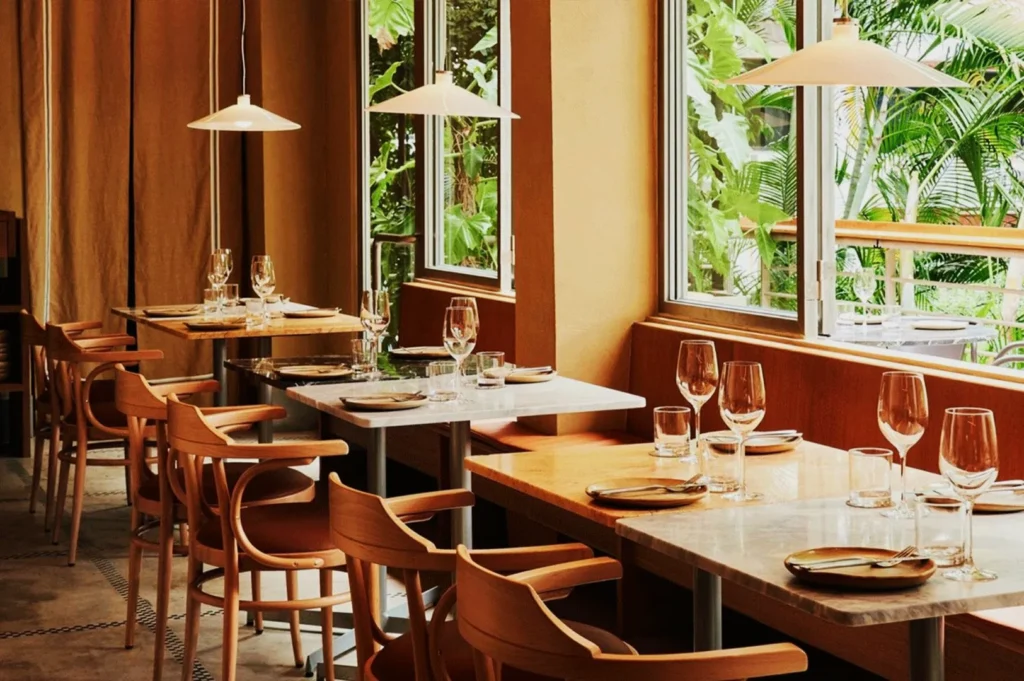 Cozy restaurant interior with wooden tables set for dining. Elegant chairs surround the tables, and large windows reveal lush greenery outside.