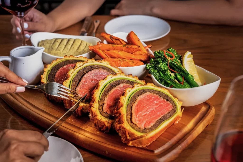 A wooden platter features sliced beef Wellington, showing juicy pink meat inside a flaky crust, with sides of roasted carrots, broccolini, and mustard.