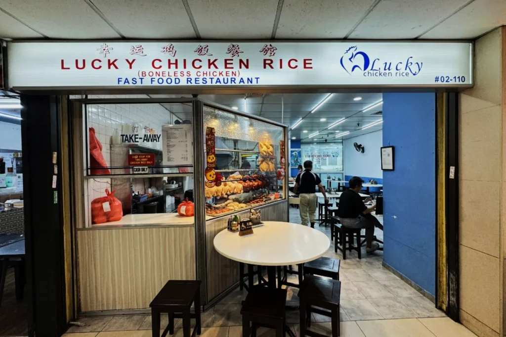 Exterior of "Lucky Chicken Rice" fast food restaurant with a display of roasted chickens. Customers are dining inside. Bright and inviting atmosphere.