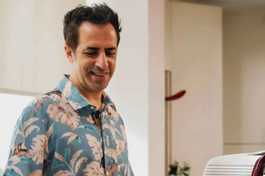 Chef Antonio Miscellaneo in a floral shirt with a relaxed expression stands indoors near a coffee machine. The setting is bright and casual, suggesting a calm atmosphere
