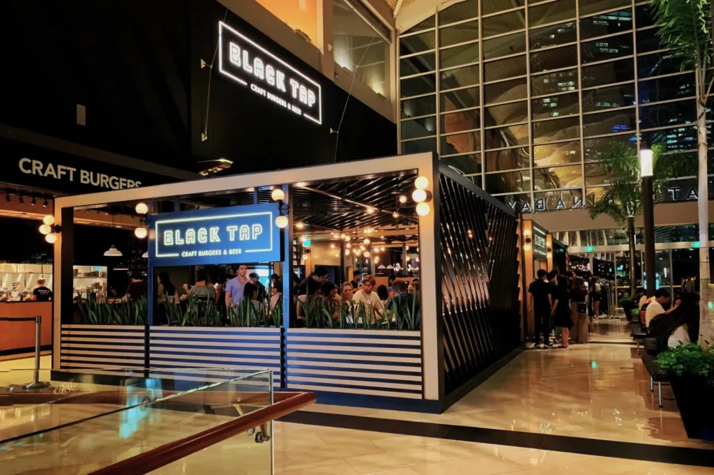 Modern restaurant, "Black Tap Craft Burgers & Beer," features a sleek black and white design, glowing lights, and a bustling, lively atmosphere.