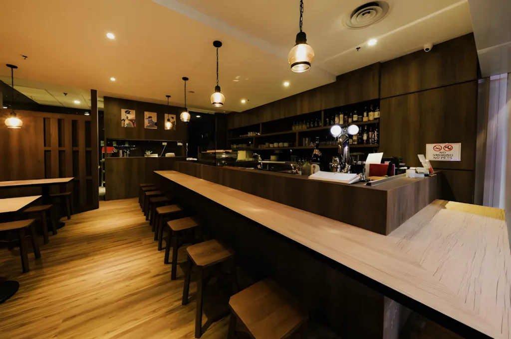 Cozy café interior with wooden decor features a long counter lined with stools, pendant lights overhead, and neatly arranged shelves in the background.