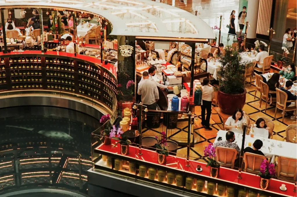 Upscale restaurant scene with elegant diners at white-clothed tables, surrounded by potted plants and warm lighting. Staff in white attire serve guests.