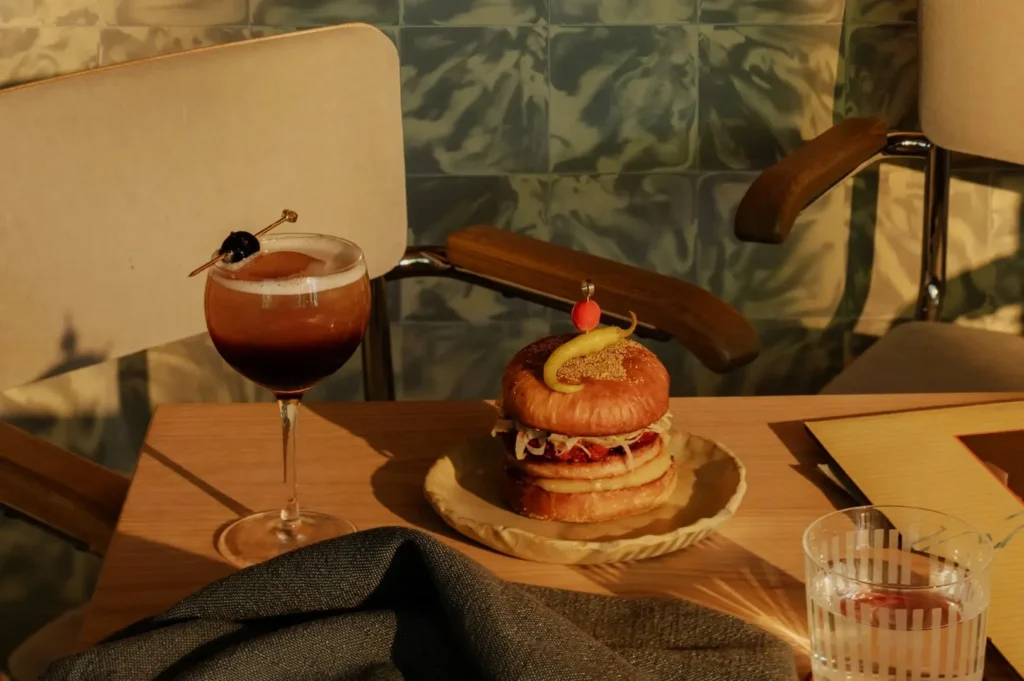 A cozy table setting features a cocktail with a garnish, a stacked sandwich on a beige plate, and a striped glass of water. Soft lighting creates a warm, inviting ambiance.
