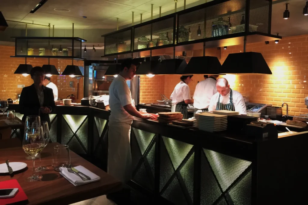 Modern restaurant kitchen with chefs in white uniforms preparing food at a sleek, black counter. Warm lighting and brick walls create a cozy ambiance.