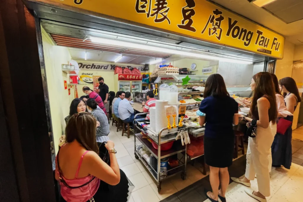 A small, bustling Yong Tau Fu stall with a yellow sign. People wait in line and sit inside, creating a lively and social atmosphere. Plates and cups are prepared on a cart.