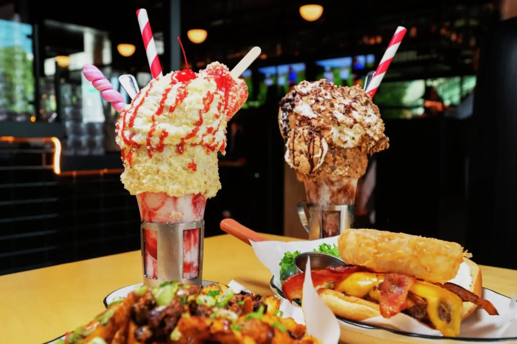Decadent milkshakes topped with whipped cream and sprinkles, alongside cheesy bacon fries and a cheeseburger. A playful, indulgent dining scene.