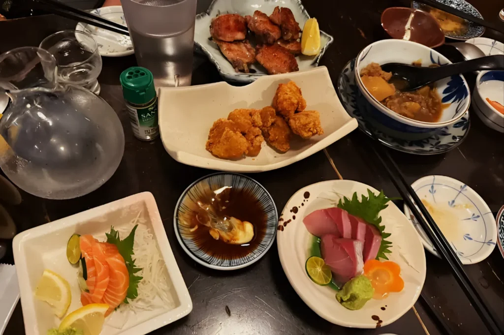 A table filled with Japanese dishes, including assorted sashimi with wasabi, fried chicken, grilled meat, soy sauce, and a jug of water. The setting is cozy and inviting.