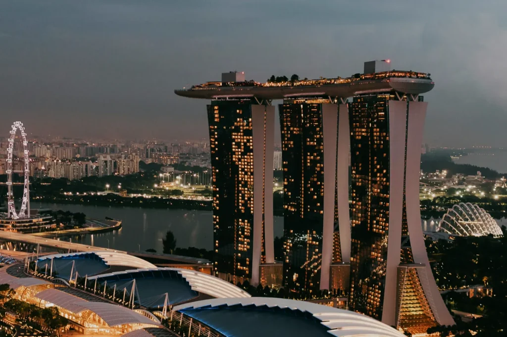 Aerial view of Marina Bay Sands at dusk in Singapore, with its illuminated towers and sky park. The Singapore Flyer and cityscape are visible in the background.