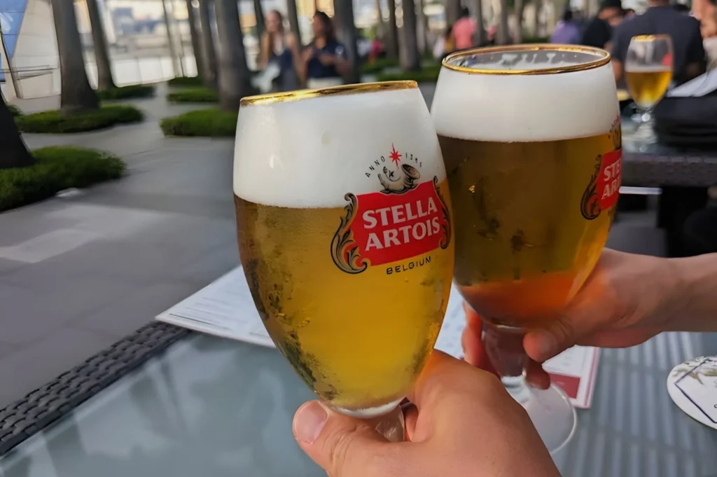 Two hands clink Stella Artois glasses filled with frothy beer on a rooftop terrace. The setting is lively, with people chatting in the background