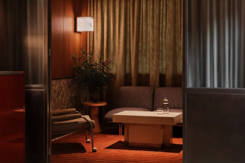 Cozy room with a modern sofa, glass-topped table with a water bottle and glass, potted plant, and soft lighting. Warm tones create a serene ambiance.
