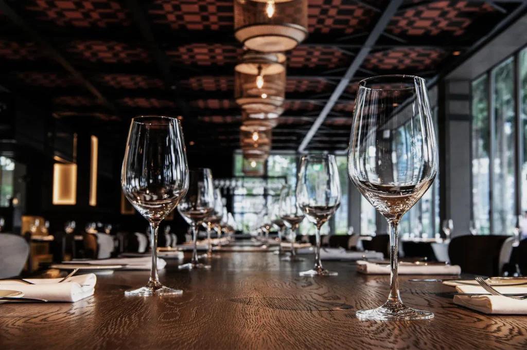 Elegant restaurant interior with a focus on a long wooden table set with polished wine glasses and folded napkins, under warm pendant lighting.