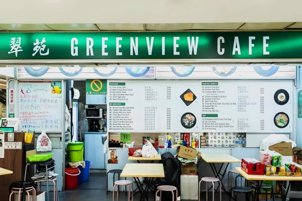 Greenview Cafe interior with a green sign above. Menu boards on the wall list various dishes. Simple tables, stools, and cartons create a casual vibe.