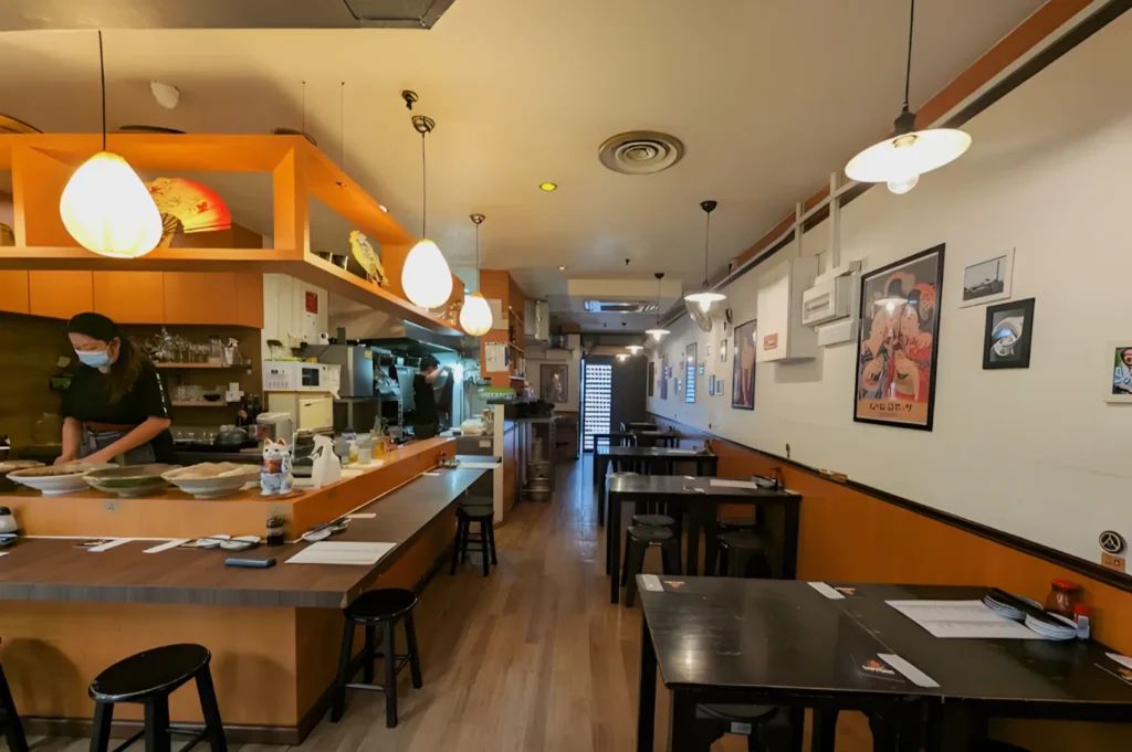 Cozy restaurant interior with warm lighting, wooden floors, and wall art. A masked staff member prepares food behind the counter, creating a peaceful ambiance.