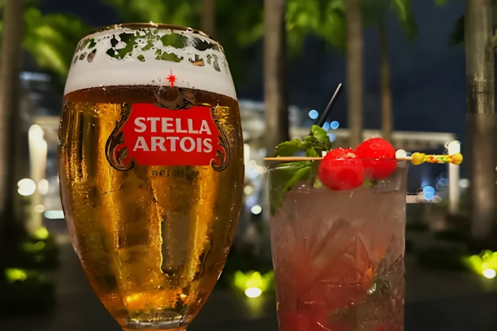 A glass of Stella Artois beer with foam beside a cocktail with cherries, ice, and garnish, set against a blurred, outdoor evening backdrop with palm trees