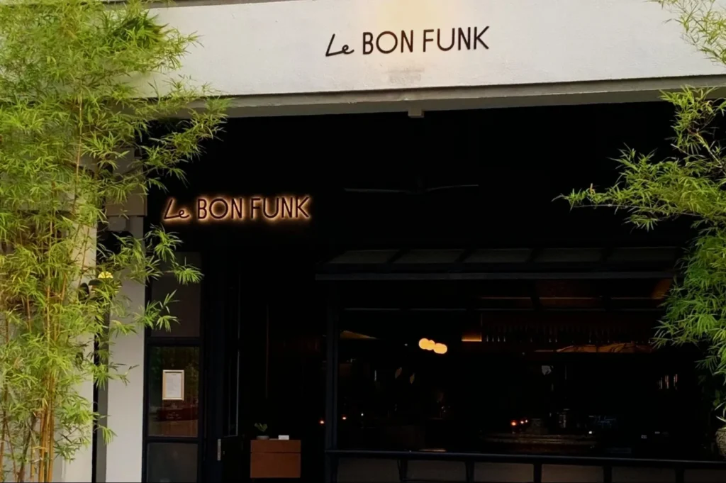 Entrance of Le Bon Funk restaurant with a minimalist exterior, featuring greenery on both sides. The neon sign radiates a warm, inviting glow.