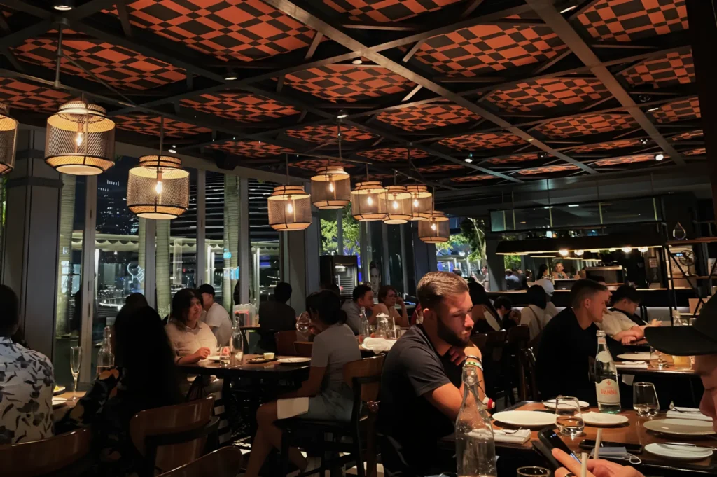 Modern restaurant interior with ambient lighting and patterned ceiling. Diners sit at wooden tables, creating a lively, cozy atmosphere.
