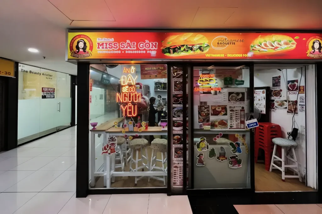 Storefront of a Vietnamese eatery named "Miss Sài Gòn" in a mall, featuring bright signage, a neon sign, interior menu displays, and stools by a counter.