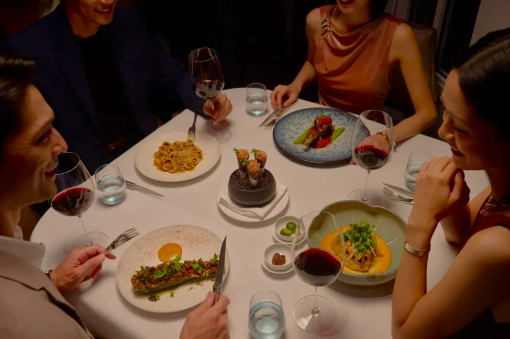 Four people enjoy an elegant dinner at a round table with gourmet dishes, red wine, and smiles, creating a warm, upscale dining ambiance.