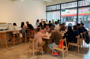 A cozy café with light wooden chairs and tables, filled with people enjoying conversations. Large windows offer a view of a passing red bus outside.