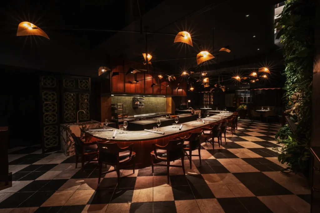 Elegant restaurant interior with curved marble counter, stylish wooden chairs, warm pendant lights, and a lush green wall, creating a cozy ambiance.