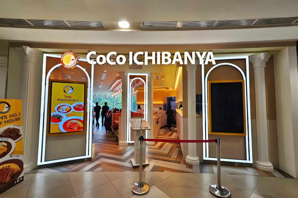 Entrance of CoCo Ichibanya restaurant with illuminated signs and yellow interior. Customers are dining inside. A menu board displays featured dishes.