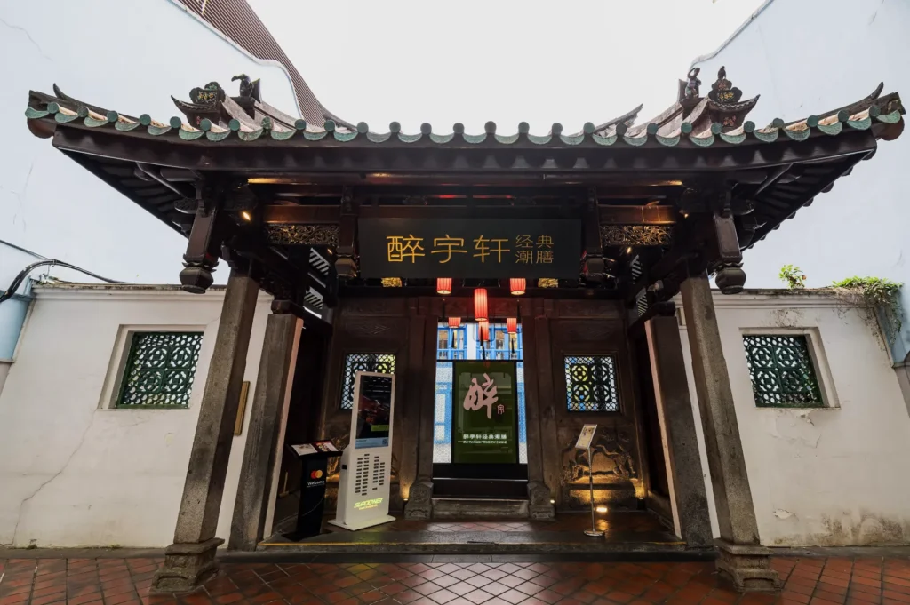 Traditional Chinese pavilion entrance with intricate woodwork, green tiled roof, and a black sign with gold characters. Lanterns hang inside, creating a welcoming atmosphere.