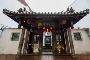 Traditional Chinese pavilion entrance with intricate woodwork, green tiled roof, and a black sign with gold characters. Lanterns hang inside, creating a welcoming atmosphere.