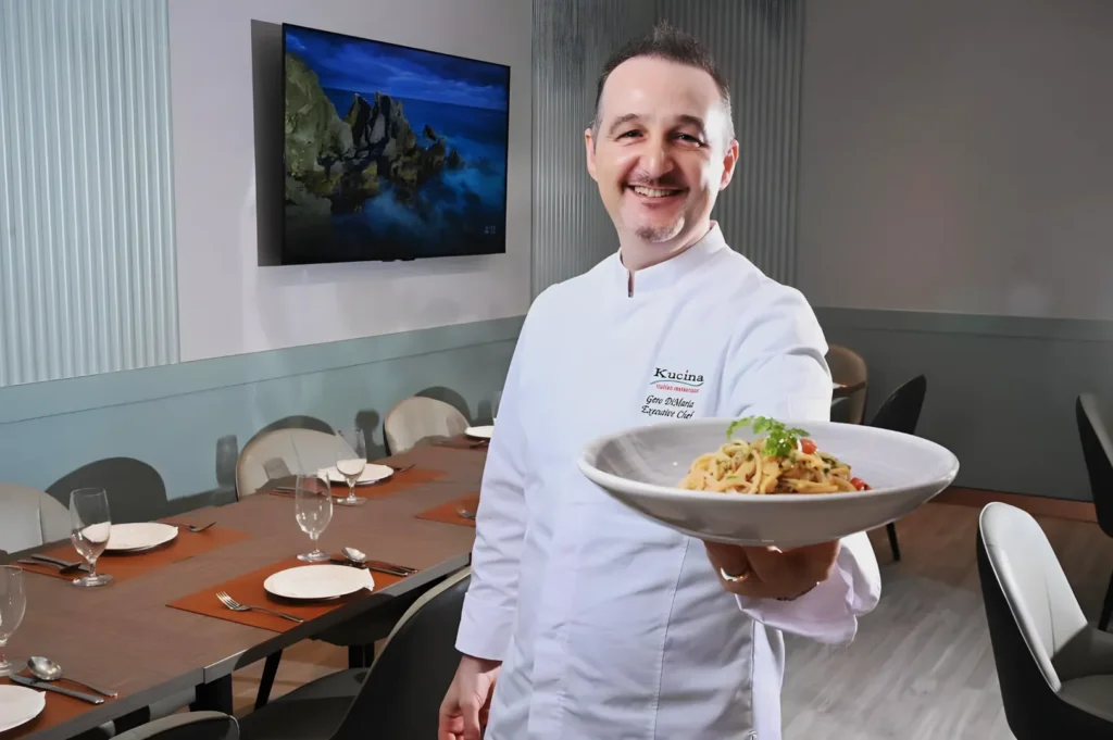 Chef in a white uniform, smiling and presenting a dish of pasta garnished with herbs in a modern dining room with a set table and a seascape on the TV.