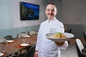 Chef in a white uniform, smiling and presenting a dish of pasta garnished with herbs in a modern dining room with a set table and a seascape on the TV.