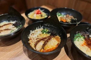 Five bowls of ramen on a wooden table, each topped with sliced pork, seaweed, sprouts, and scallions, creating a warm, inviting atmosphere.