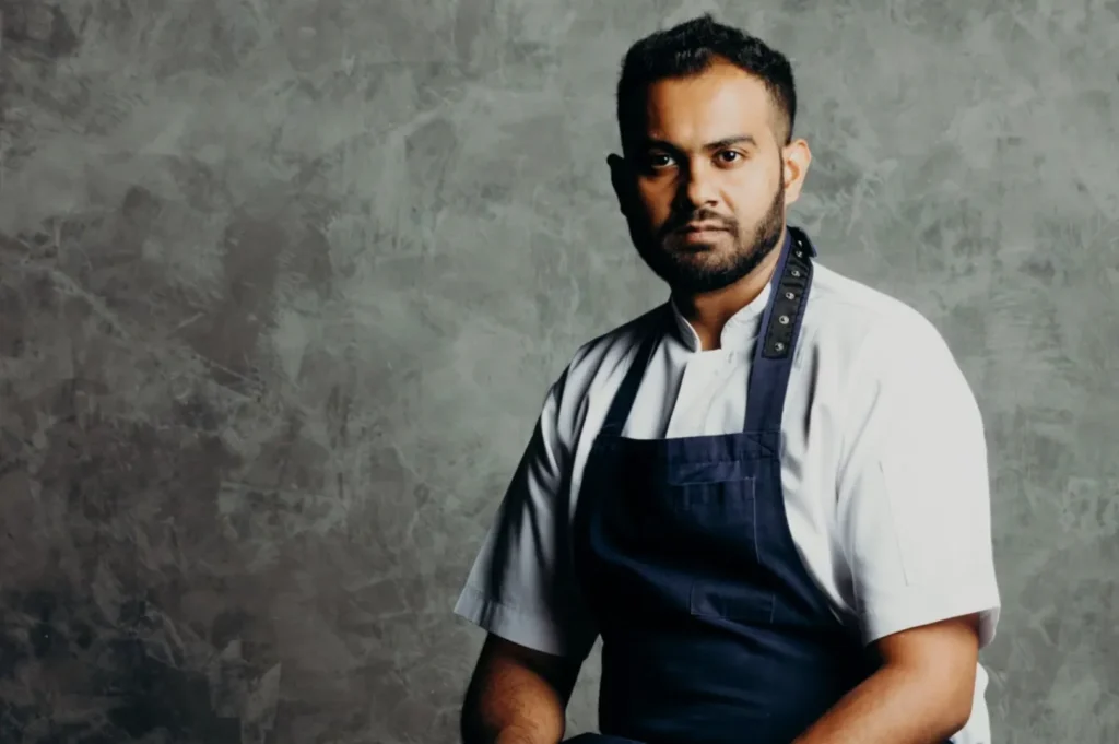 A chef wearing a white shirt and dark apron stands against a textured gray background. He looks confidently at the camera, conveying professionalism.