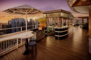 Elegant rooftop bar at sunset with wooden floors, sleek bar stools, and striped umbrellas. Skyscrapers in the background create a vibrant cityscape.