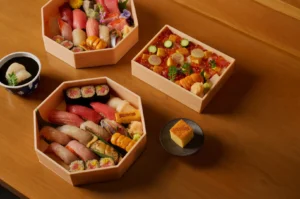 Assorted sushi and sashimi in wooden boxes on a wooden table, accompanied by a cup of tea and a slice of tamagoyaki, creating a traditional Japanese dining scene.