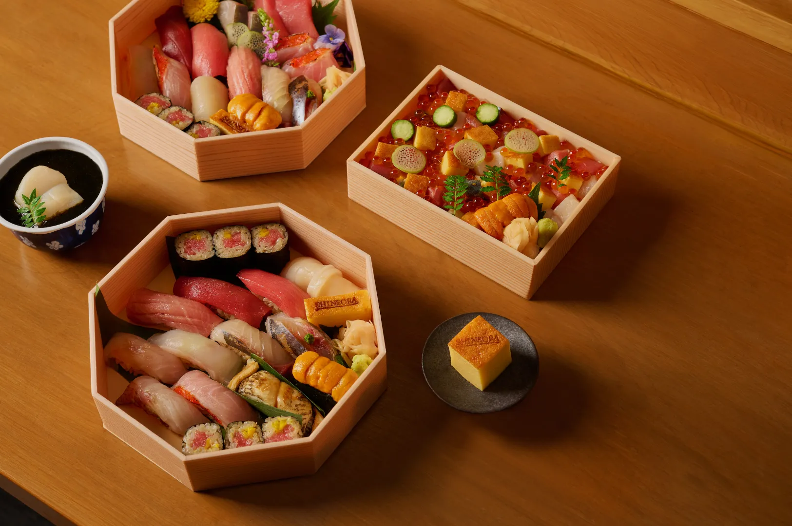 Assorted sushi and sashimi in wooden boxes on a wooden table, accompanied by a cup of tea and a slice of tamagoyaki, creating a traditional Japanese dining scene.