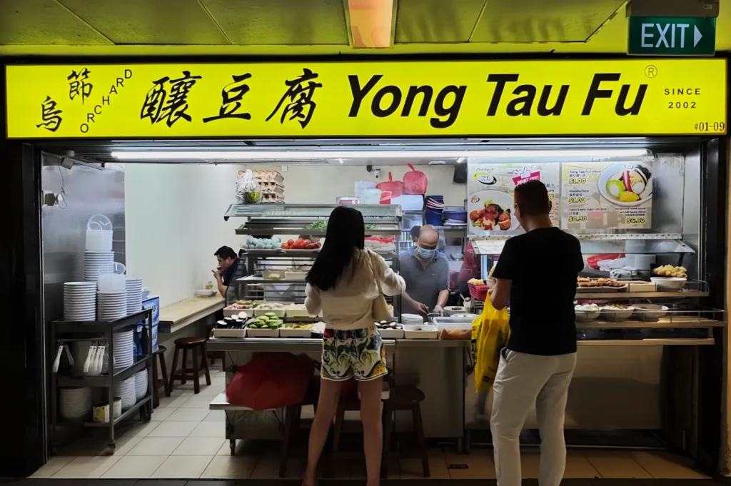 A yellow sign reads "Yong Tau Fu" at a food stall. Two customers are selecting ingredients from a variety of fresh options, while a masked staff member prepares food behind the counter. The setting has a lively, casual atmosphere.