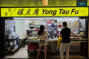 A yellow sign reads "Yong Tau Fu" at a food stall. Two customers are selecting ingredients from a variety of fresh options, while a masked staff member prepares food behind the counter. The setting has a lively, casual atmosphere.
